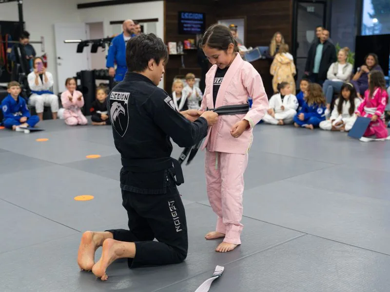 Coach awarding a new belt to a young student at Legion while parents and classmates watch