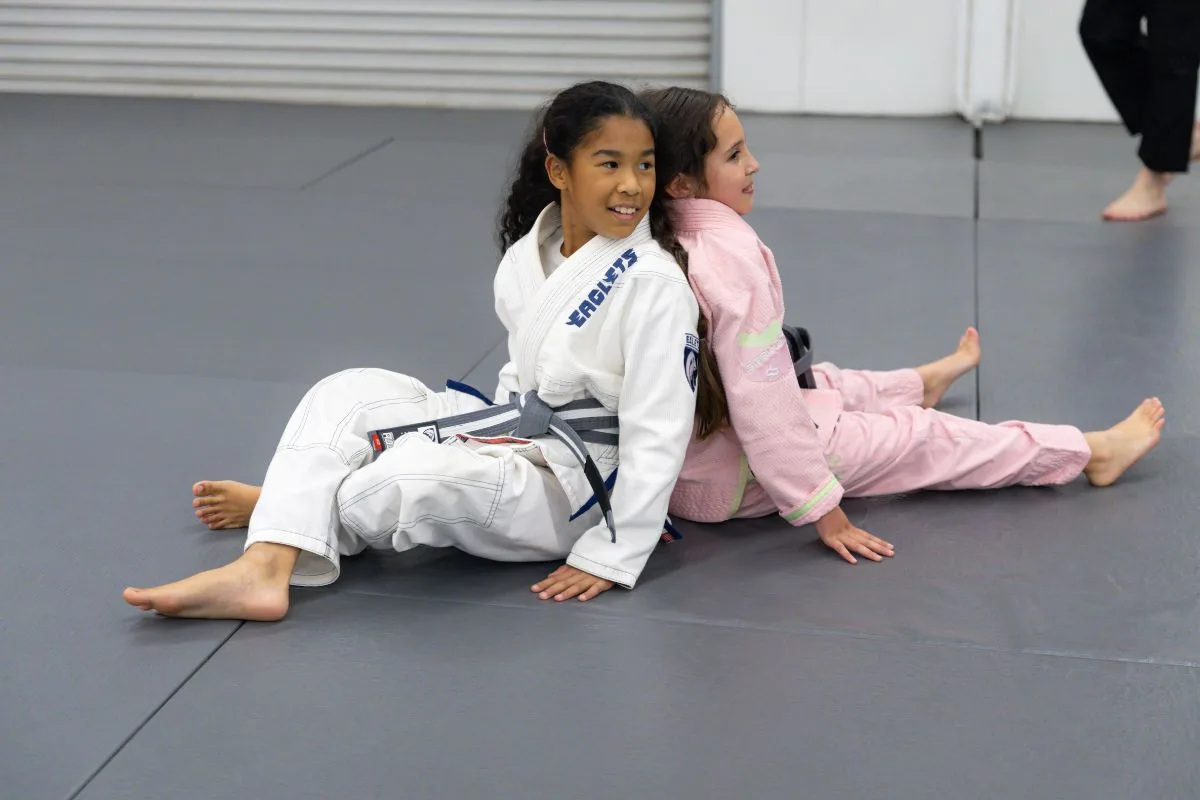 Two smiling girls in gis sitting back to back on the Legion mats